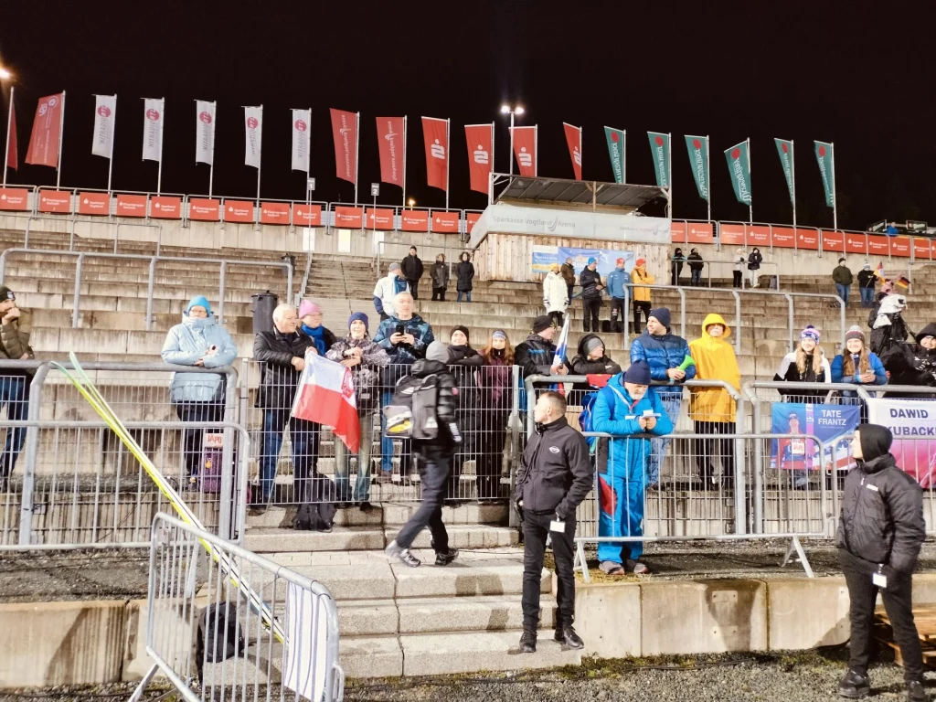 Polscy kibice w Klingenthal z Kacprem Tomasiakiem Grupa kibiców ubranych w zimowe ubrania stoi na trybunach stadionu, część z nich trzyma flagę Polski, a w tle widoczne są puste rzędy, liczne flagi oraz banery. Oświetlenie sugeruje, że wydarzenie odbywa się wieczorem.