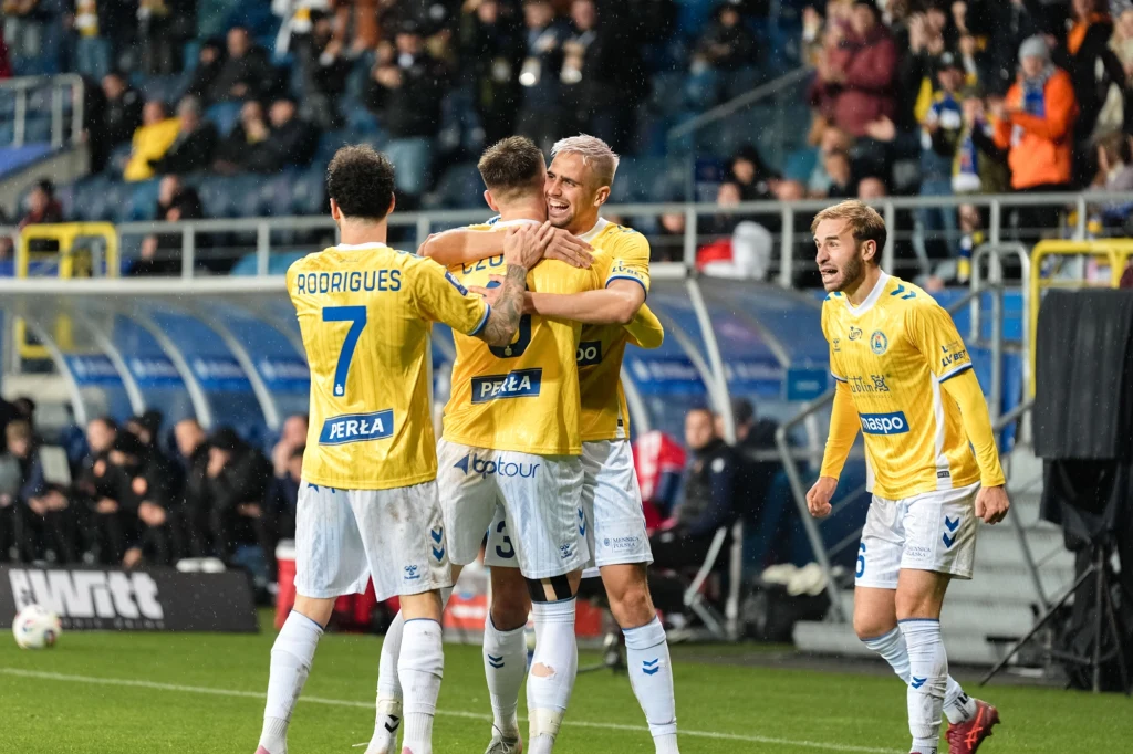 Piłkarze w żółtych strojach świętują zdobycie bramki na boisku piłkarskim, otoczeni przez kibiców na trybunach.