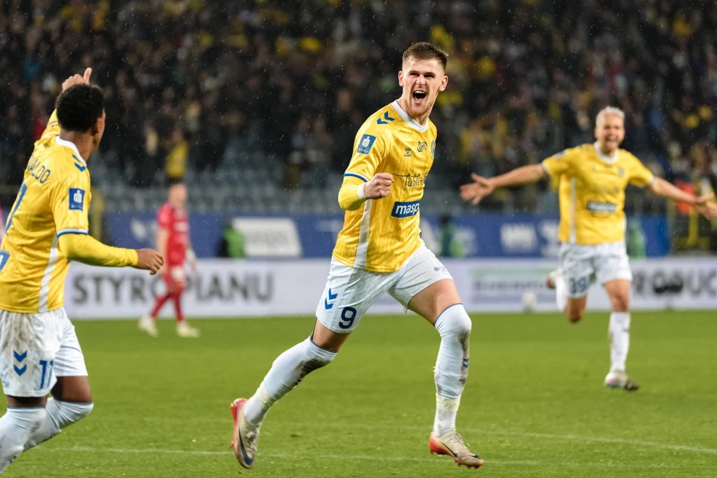 Piłkarz w żółtej koszulce świętuje zdobycie gola na stadionie, z dwoma innymi zawodnikami podbiegającymi z uniesionymi rękami w tle, tłum kibiców na trybunach.