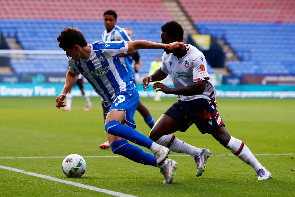 Dwóch piłkarzy rywalizuje o piłkę na murawie stadionu, jeden zawodnik próbuje wyprzedzić drugiego, zbliżając się do linii bocznej boiska, w tle rozmyci zawodnicy i kolorowe trybuny.