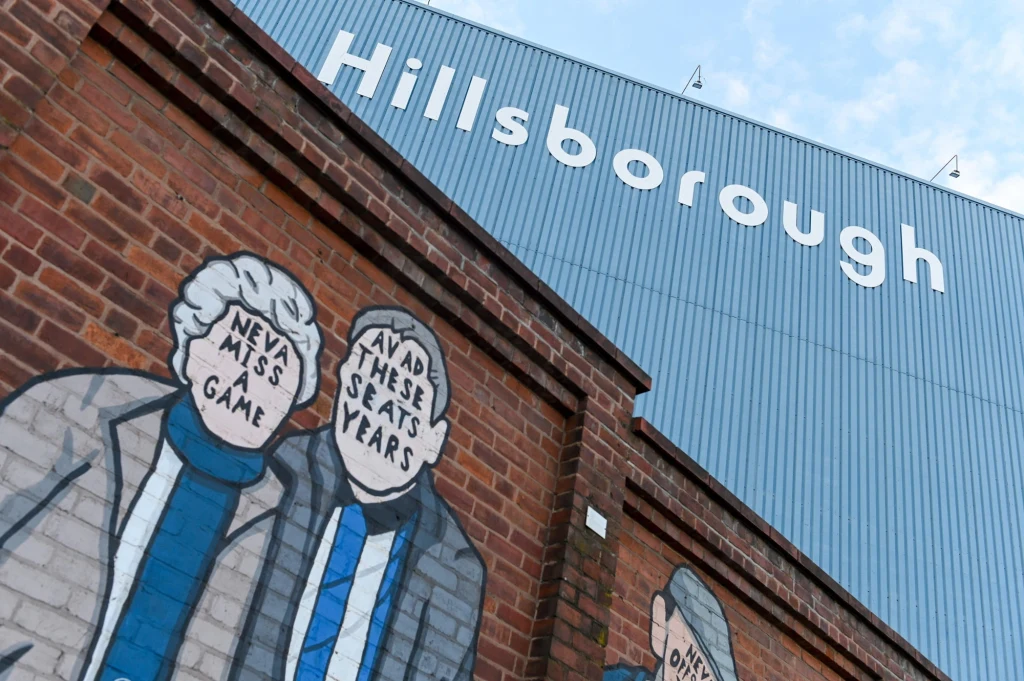 Fragment murala ściennego z wizerunkami osób o niewyraźnych, opisowych twarzach, na tle stadionu piłkarskiego Hillsborough. Budynek stadionu i jego nazwa dominują nad sceną, podkreślając sportowy i kulturowy charakter miejsca.