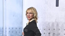 Elegancko ubrana kobieta w czarnej sukience i czarnych szpilkach pozuje bokiem, stojąc na szarej podłodze na tle dekoracyjnym przypominającym metalowe nity oraz morze. Oświetlenie podkreśla jej sylwetkę i blond włosy.