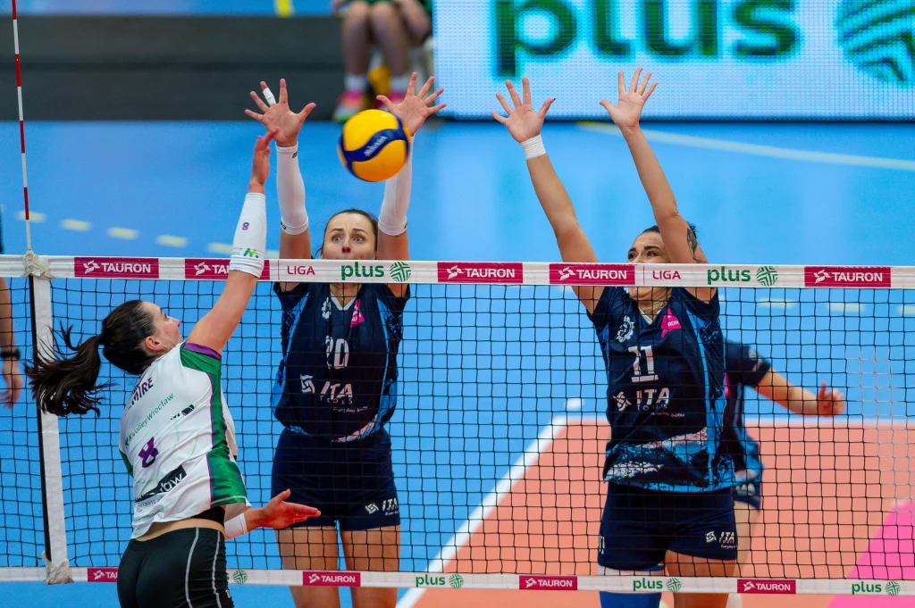 Starcie #Volley Wrocław ze Stalą Mielec musiał rozstrzygnąć tie-break Zawodniczka drużyny w białym stroju atakuje piłkę siatkarską, próbując przebić ją przez siatkę, podczas gdy dwie zawodniczki w ciemnych strojach wykonują blok obiema rękami, starając się zatrzymać przeciwniczkę. W tle widoczna jest część boiska oraz si...