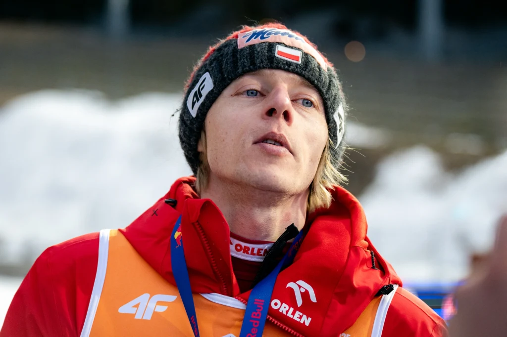 Dawid Kubacki Młody mężczyzna w sportowym stroju zimowym, ubraną w czerwoną kurtkę, czapkę z logo oraz polską flagą na froncie. Na szyi wisi medal lub plakietka. W tle rozmyte śnieżne otoczenie.