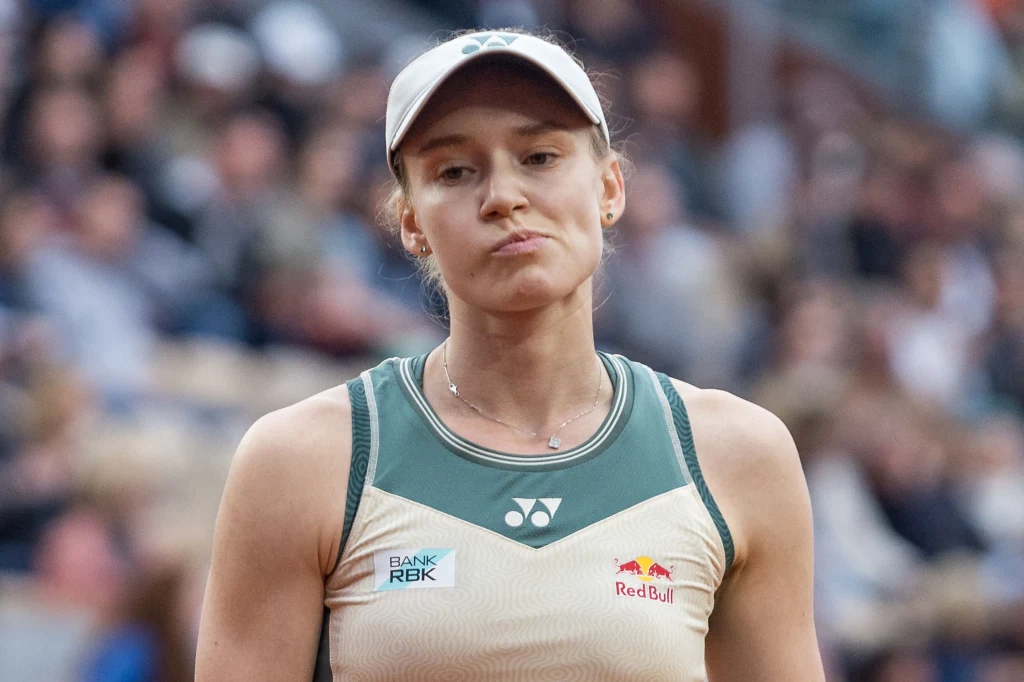 Tennisistka w czapce z daszkiem w stroju sportowym, z widocznymi logotypami sponsorów na koszulce, stoi na tle rozmytej publiczności z wyrazem zamyślenia lub rozczarowania na twarzy.