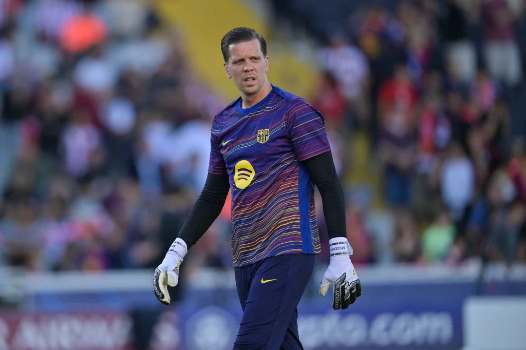 Bramkarz w sportowym stroju treningowym z logo drużyny FC Barcelona oraz logotypem sponsora na koszulce, stojący na tle zamazanego stadionu z widownią w tle.