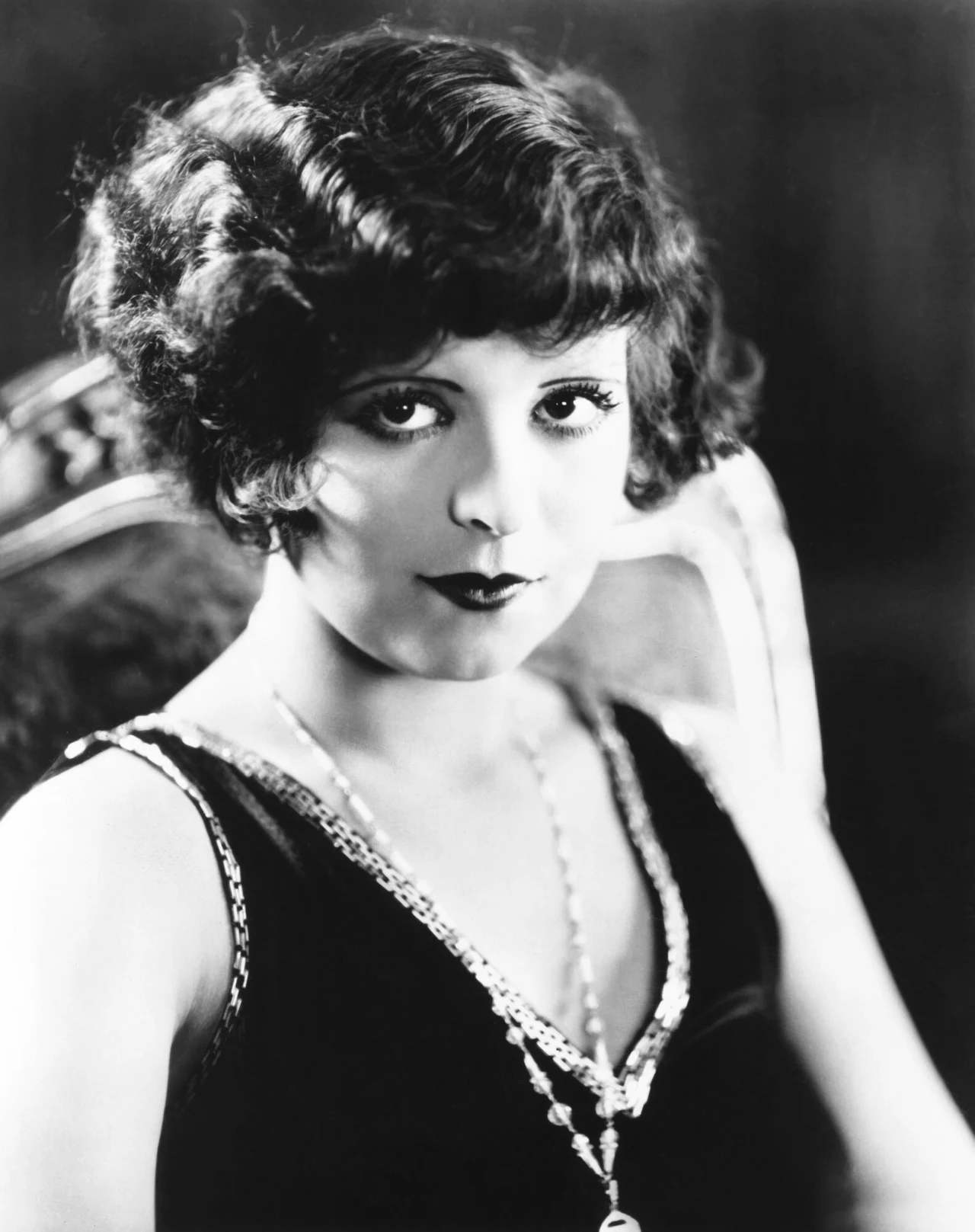 Portret młodej kobiety w stylu retro, z krótkimi, falującymi włosami, wyrazistym makijażem i elegancką biżuterią, siedzącej na ozdobnym fotelu, tło rozmyte i ciemne.