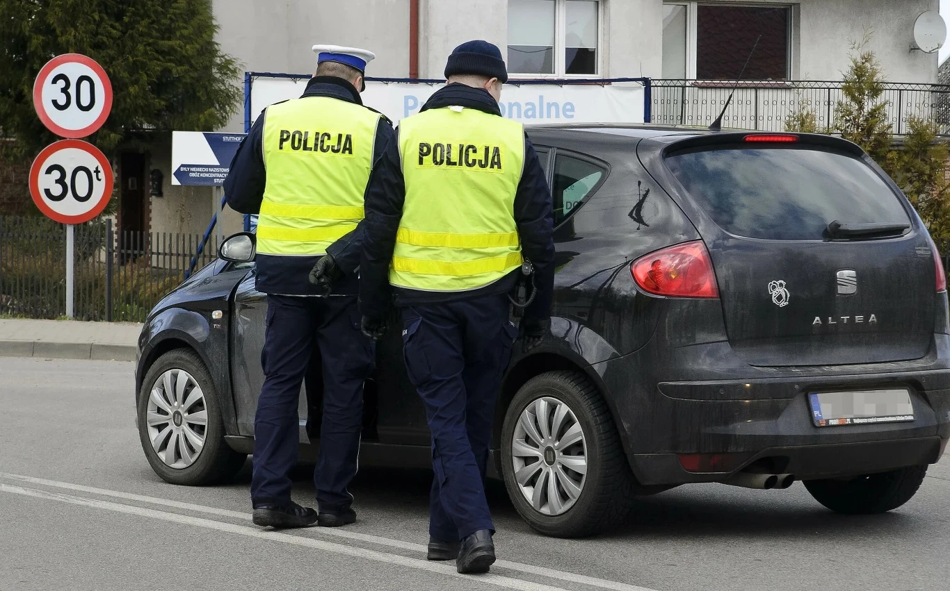 Za niewłaściwe oświetlenie pojazdu policjant może wystawić mandat 3000 zł i zatrzymać dowód rejestracyjny Dwóch policjantów w odblaskowych kamizelkach kontroluje kierowcę czarnego samochodu osobowego, pojazd zatrzymany na ulicy obok znaków ograniczenia prędkości do 30 km/h.