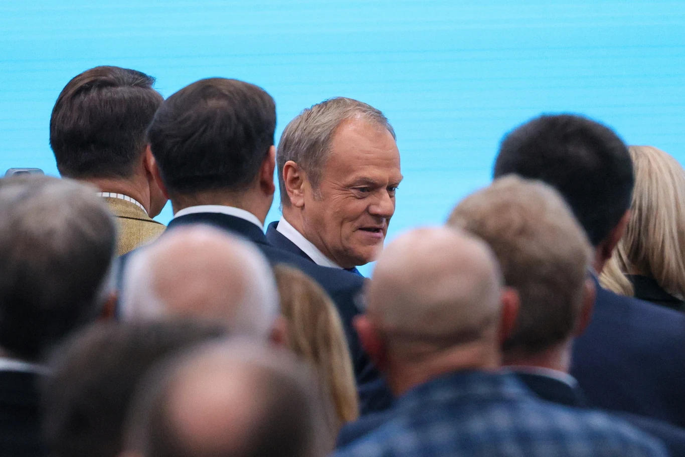 Premier Donald Tusk tuż po przemówieniu na kongresie zjednoczeniowym Mężczyzna w garniturze uśmiechający się wśród licznej grupy ludzi, zwróconych do niego plecami, na jasnoniebieskim tle.