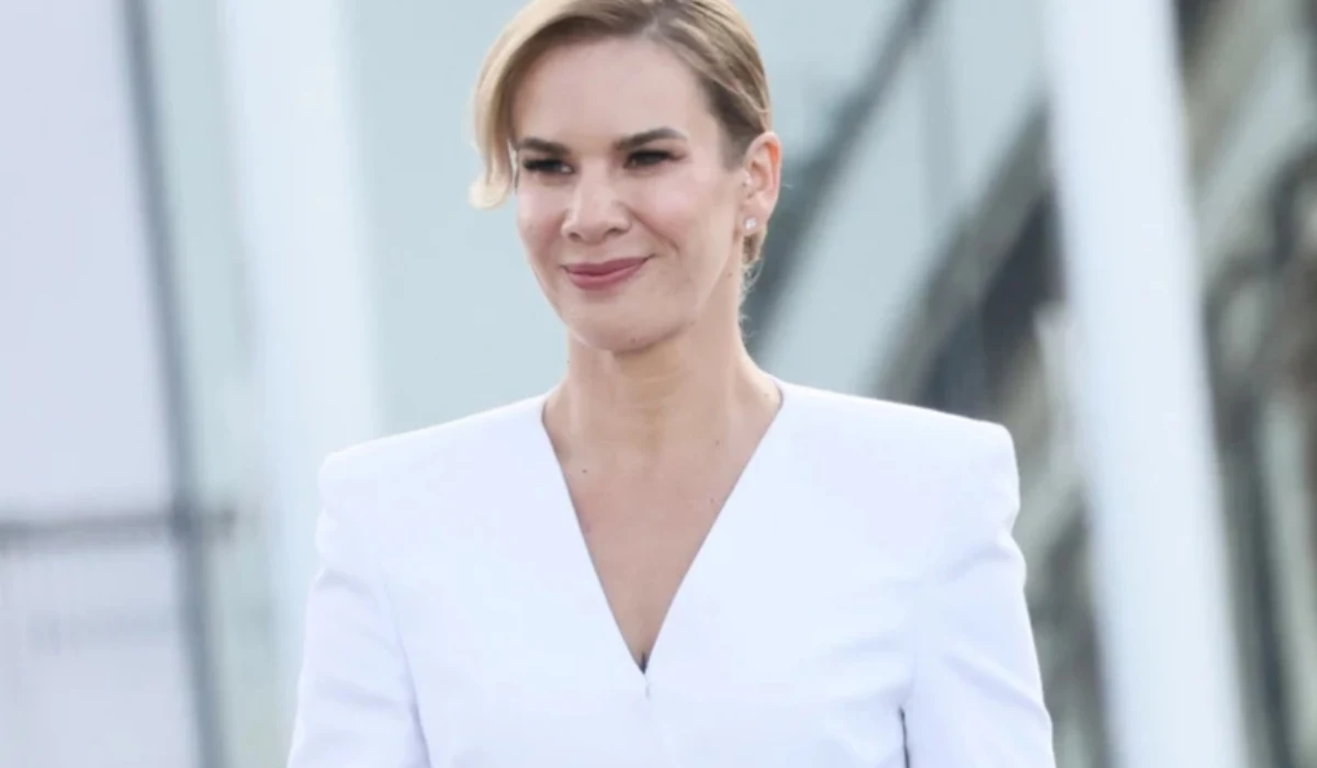 Marta Nawrocka Elegancka kobieta w białym żakiecie z delikatnym uśmiechem, tło rozmyte i nowoczesne, włosy spięte, subtelny makijaż, kompozycja skupia się na osobie.