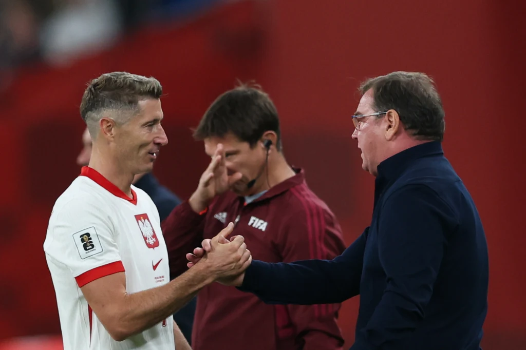 Dwóch mężczyzn wymienia uścisk dłoni na tle czerwonej ściany, jeden z nich ubrany w biało-czerwoną koszulkę piłkarską z godłem Polski, drugi w ciemnej marynarce, w tle osoba w sportowym stroju z plakietką FIFA na piersi.