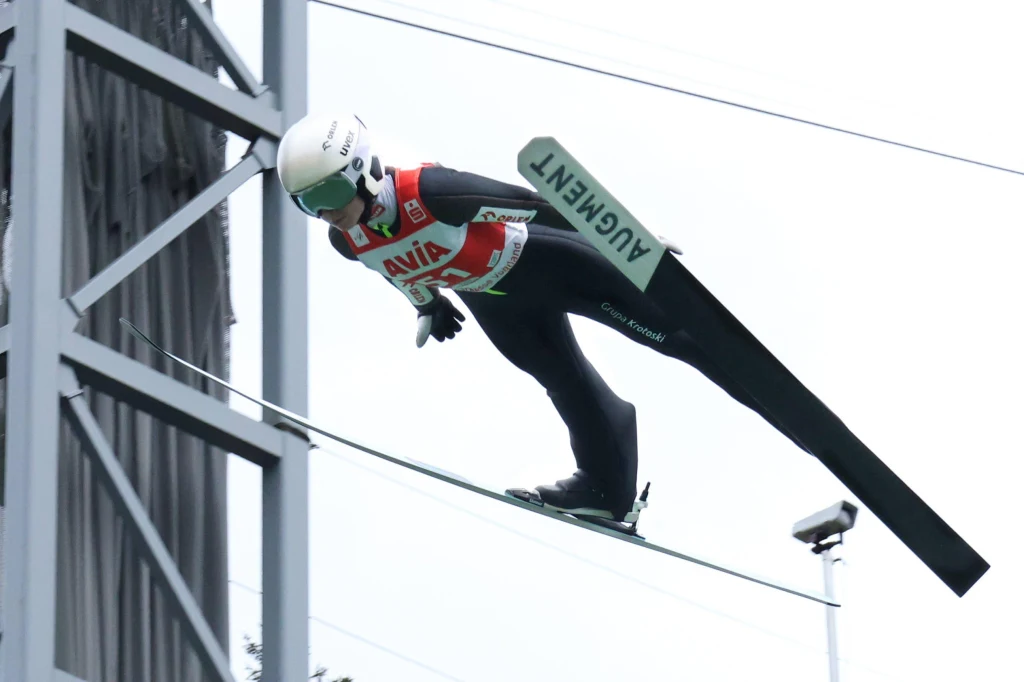 Anna Twardosz w Klingenthal Zawodnik w stroju narciarskim w trakcie lotu podczas konkursu skoków narciarskich, w pełnym wyposażeniu ochronnym, w tle fragment konstrukcji skoczni.