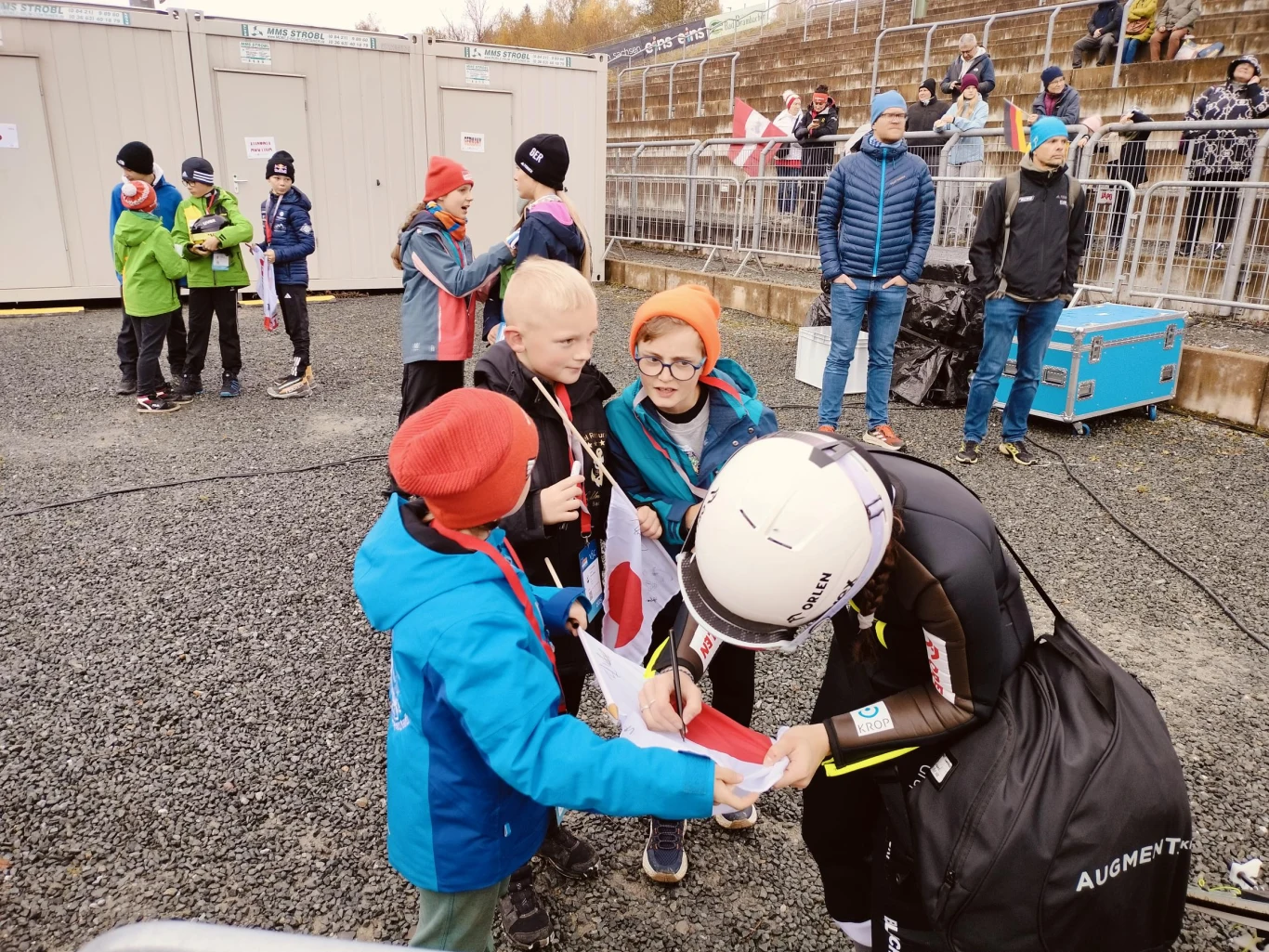 Sportowiec w stroju startowym podpisuje autograf dzieciom ubranym w kolorowe kurtki i czapki, wokół stoją inni ludzie na tle metalowych barierek i kontenerów technicznych.