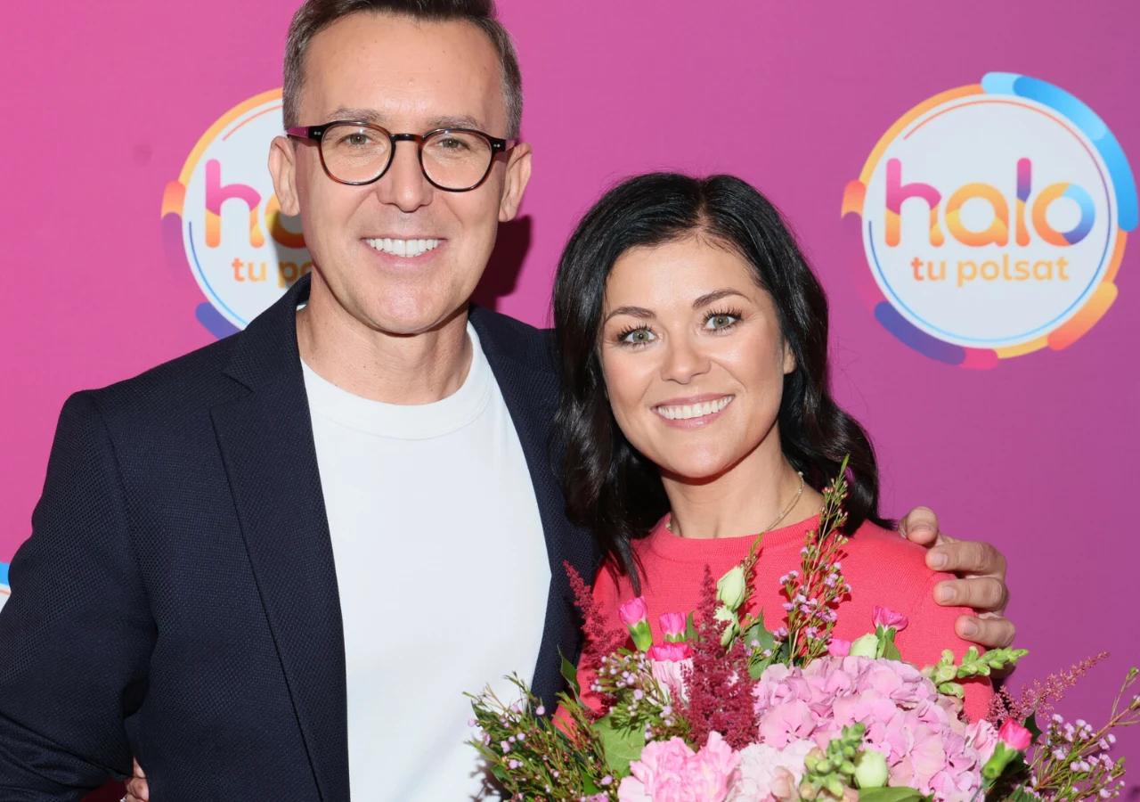 Katarzyna Cichopek i Maciej Kurzajewski Dwie uśmiechnięte osoby, jedna z nich trzyma bukiet kwiatów, pozują na tle różowej ścianki z logo „halo tu Polsat”.