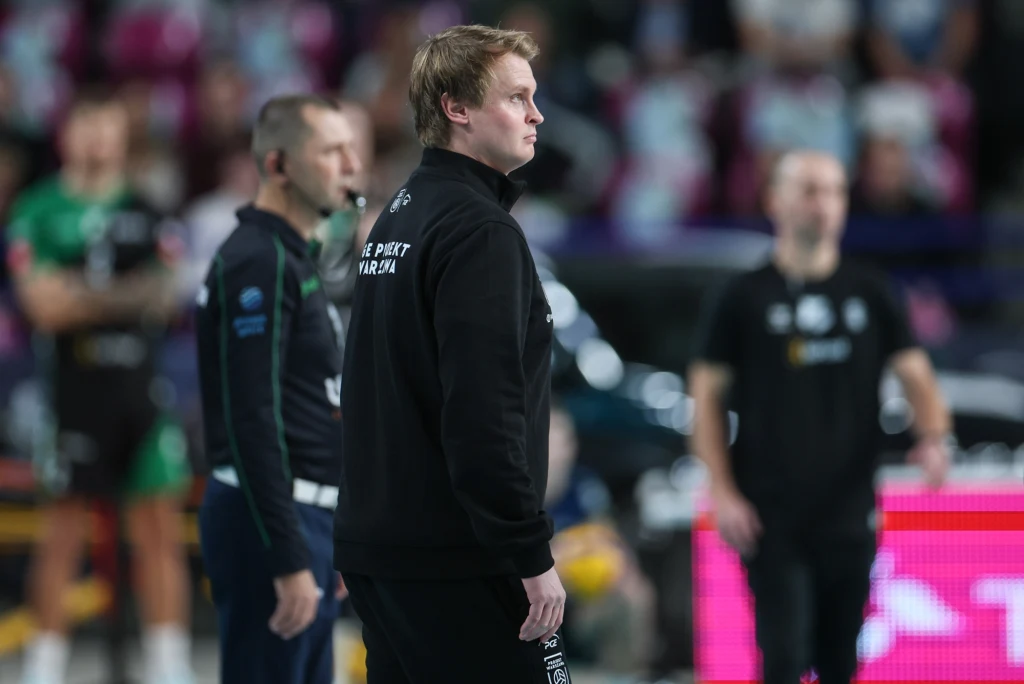 Trener PGE Projektu Warszawa Tommi Tilikainen Trener drużyny sportowej w czarnej bluzie i spodniach stoi na hali podczas meczu, wokół niego znajdują się inni członkowie sztabu i sportowcy, tło stanowi rozmyta publiczność i kolorowe światła.