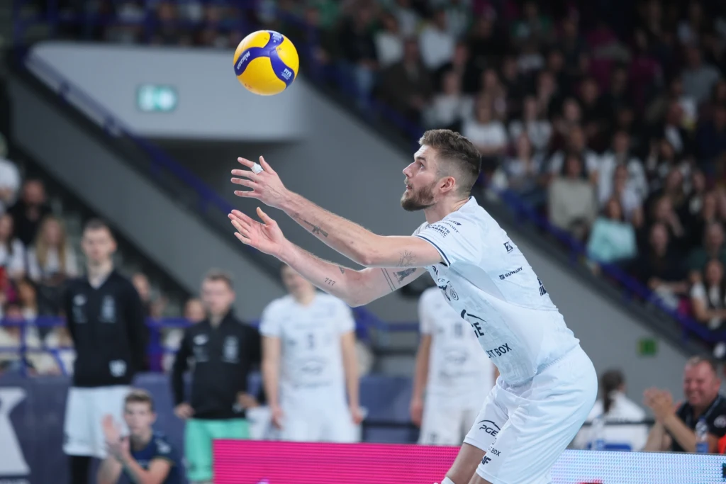 Karol Kłos, nowy środkowy PGE Projektu Warszawa Siatkarz w białym stroju wykonuje odbiór piłki podczas meczu halowego, w tle widoczni inni zawodnicy oraz publiczność.
