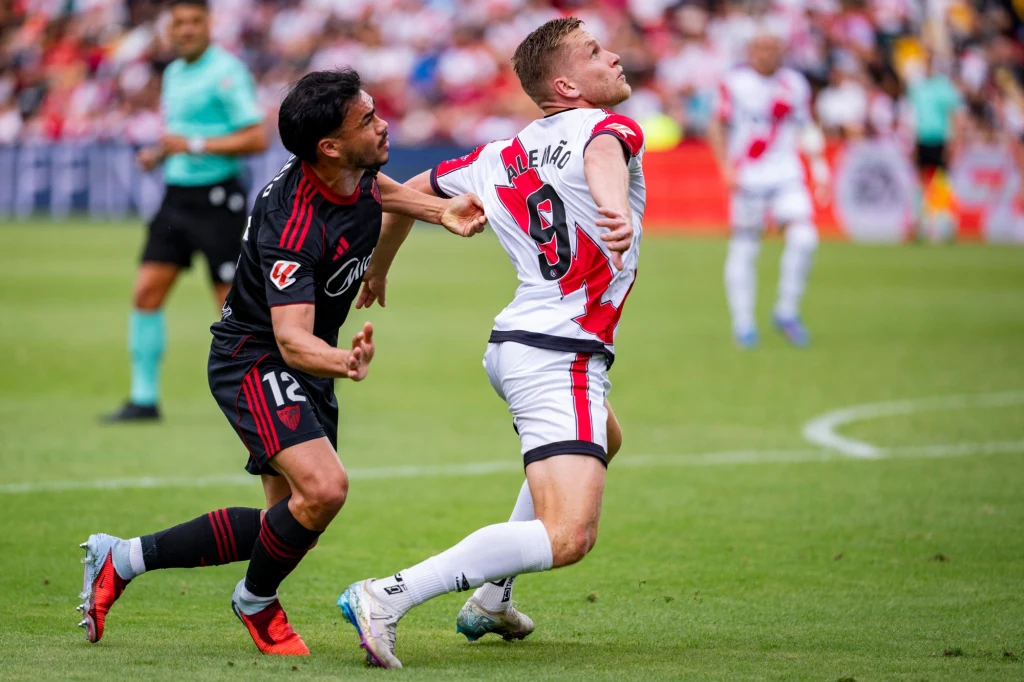 Alemao Dwóch piłkarzy walczy o piłkę podczas meczu, jeden z nich próbuje zablokować przeciwnika poprzez fizyczny kontakt, na tle rozmytej publiczności i innych zawodników na murawie.