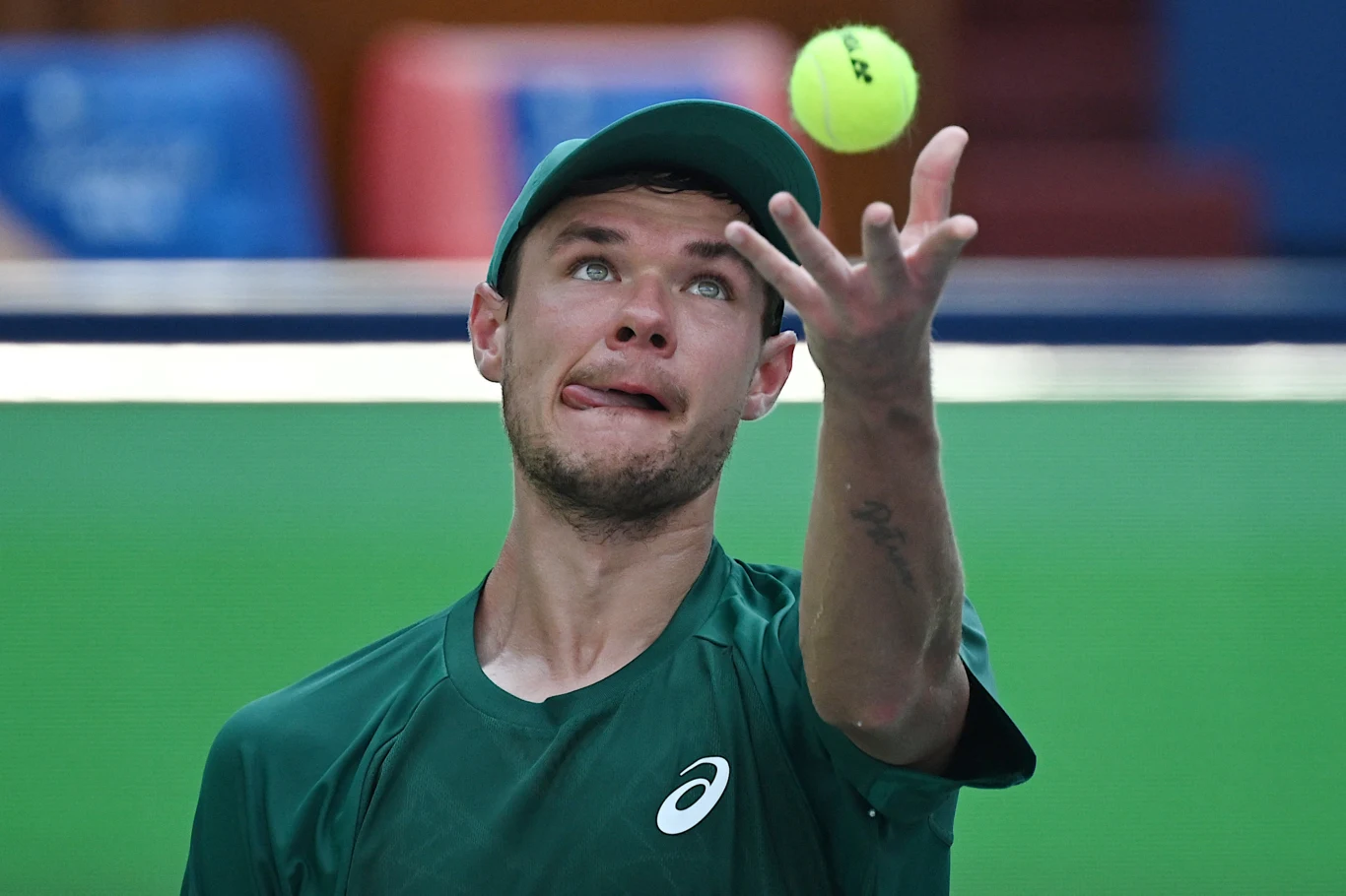 Mężczyzna w zielonej koszulce sportowej i czapce rzuca żółtą piłkę tenisową, koncentrując się na serwisie podczas meczu tenisowego.