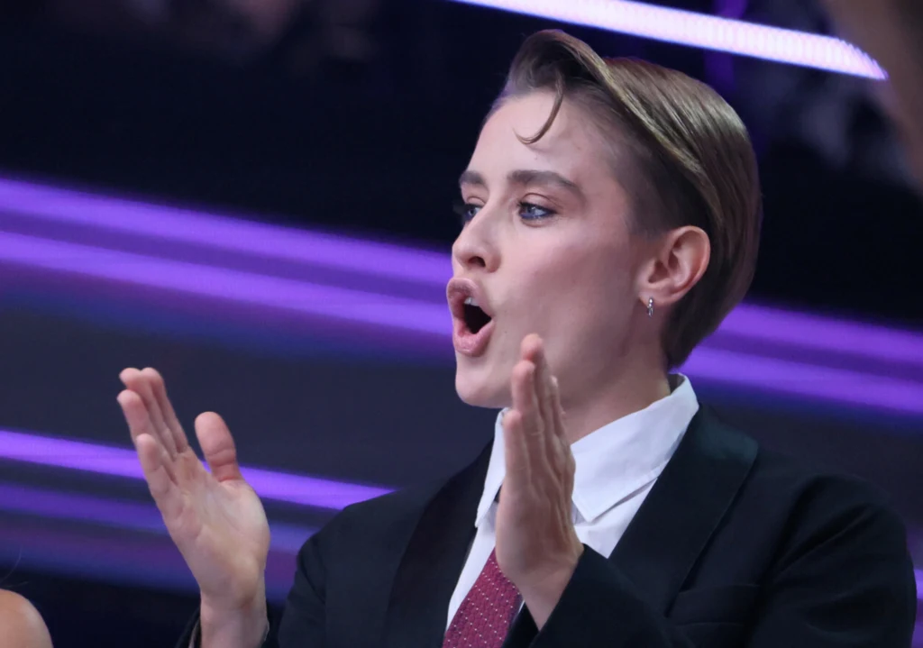 Osoba w garniturze i krawacie z krótkimi włosami, wyraźnie zaangażowana emocjonalnie, klaszcze w dłonie i ma otwarte usta jakby mówiła lub krzyczała. Za nią widoczne fioletowe światła tworzące efektowne tło.