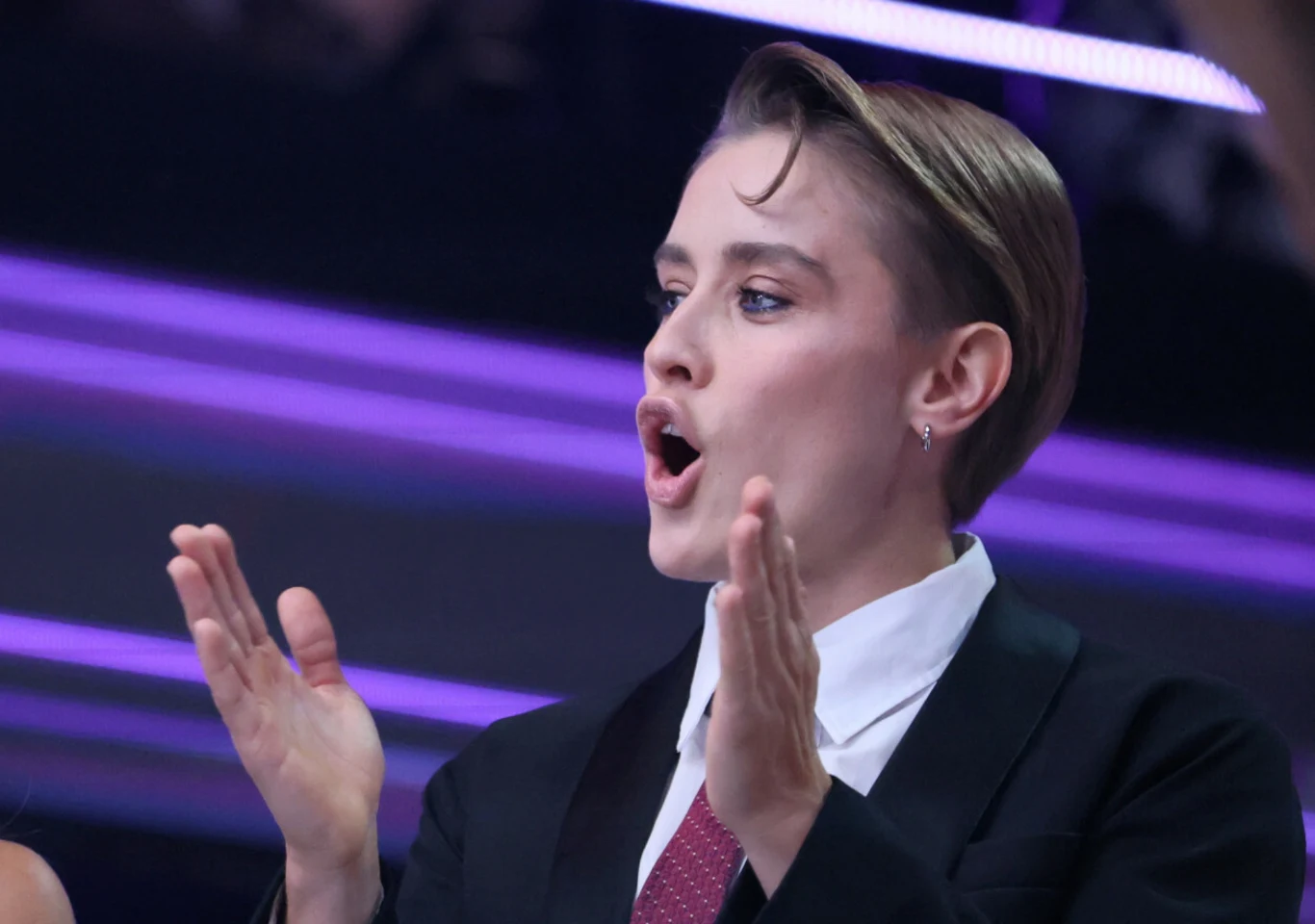 Katarzyna Zillmann w programie "Taniec z Gwiazdami" Osoba w garniturze i krawacie z krótkimi włosami, wyraźnie zaangażowana emocjonalnie, klaszcze w dłonie i ma otwarte usta jakby mówiła lub krzyczała. Za nią widoczne fioletowe światła tworzące efektowne tło.