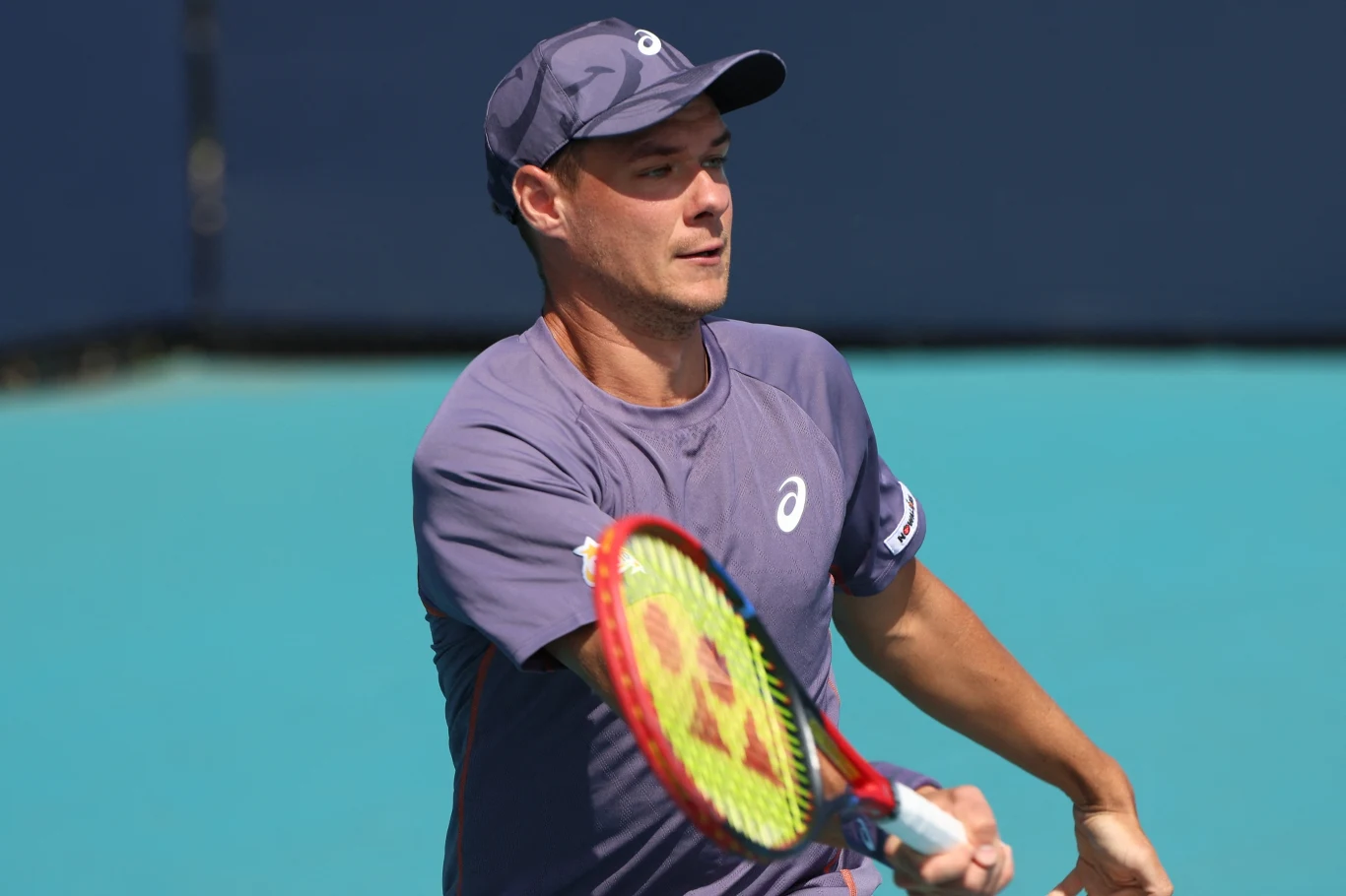 Mężczyzna w czapce sportowej podczas gry w tenisa, skupiony na odbiciu piłki rakietą, ubrany w fioletową koszulkę i znajdujący się na tle niebieskiego kortu tenisowego.