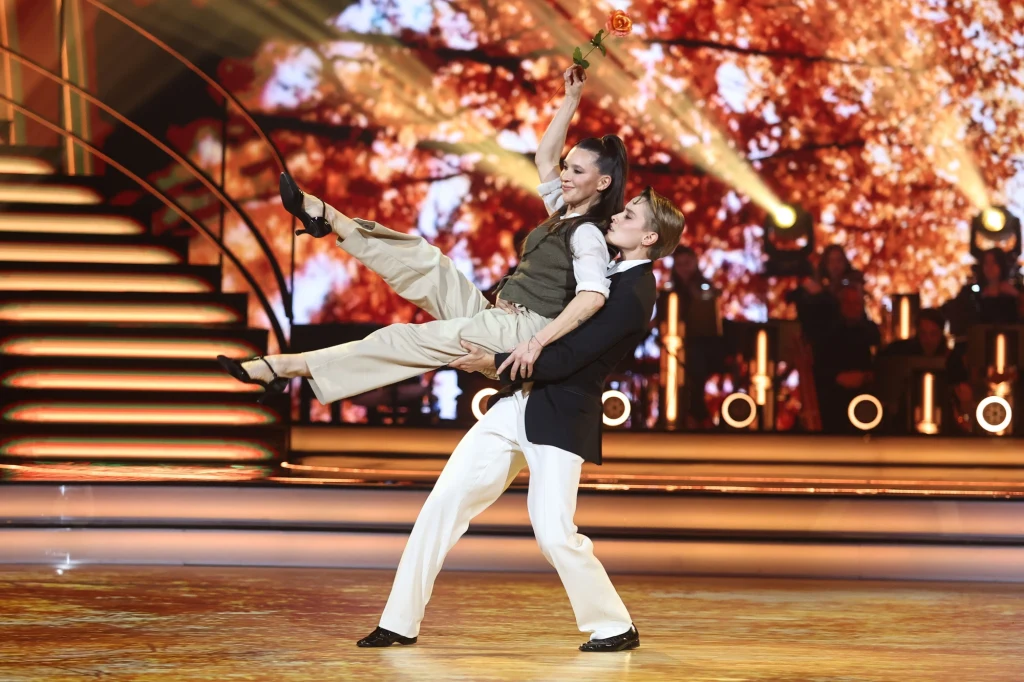 Katarzyna Zillmann i Janja Lesar w "Tańcu z Gwiazdami" Dwoje tancerzy w eleganckich strojach wykonuje dynamiczny układ taneczny na jasno oświetlonej scenie. W tle widoczne są rozświetlone schody oraz dekoracja w ciepłych barwach, a całość otoczona jest efektownym oświetleniem.