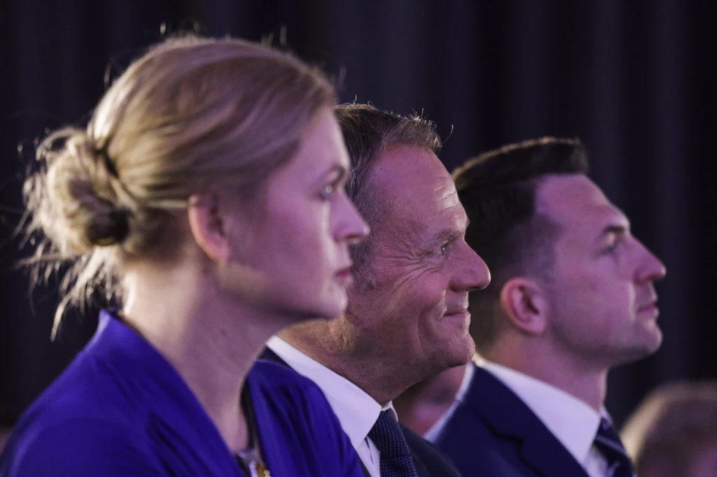 Od lewej: Barbara Nowacka (przewodnicząca Inicjatywy Polska), Donald Tusk (przewodniczący Platformy Obywatelskiej), Adam Szłapka (przewodniczący Nowoczesnej) Trzy osoby w eleganckich strojach siedzące obok siebie w skupieniu, dwie postaci patrzą przed siebie, mężczyzna w środku delikatnie się uśmiecha, tło ciemne i rozmyte podkreślające głównych bohaterów.