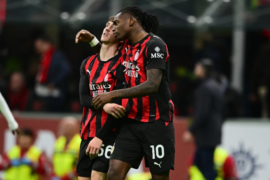 Dwóch piłkarzy drużyny AC Milan w czarno-czerwonych strojach ściska się na boisku, jeden obejmuje drugiego ramieniem, w tle rozmyty obraz trybun z kibicami.