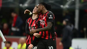 Dwóch piłkarzy drużyny AC Milan w czarno-czerwonych strojach ściska się na boisku, jeden obejmuje drugiego ramieniem, w tle rozmyty obraz trybun z kibicami.