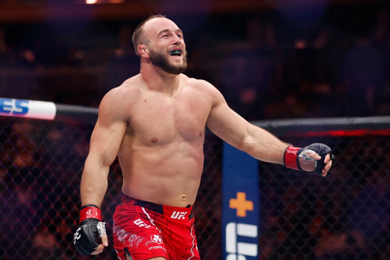 Zawodnik MMA w czerwonych spodenkach stoi w oktagonie, uśmiechnięty, z uniesioną ręką. Otoczenie wskazuje na walkę sportową, a atmosfera jest dynamiczna.