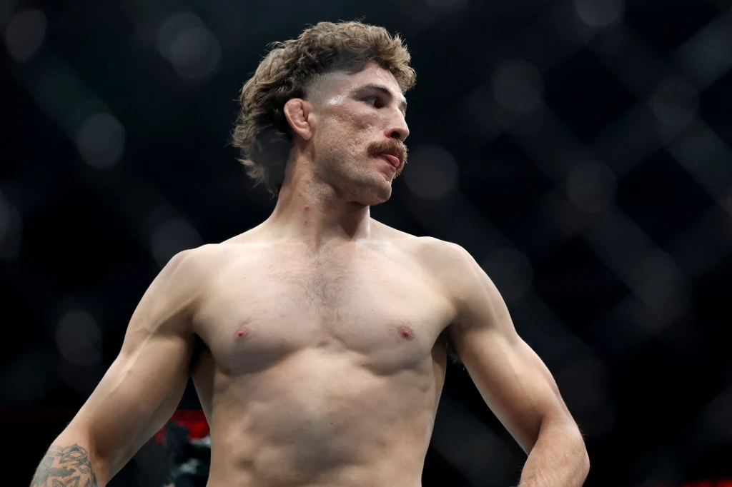 Quillan Salkilld Sportowiec z widocznymi obrażeniami na twarzy, stojący bez koszulki na arenie walki, w tle widoczna siatka klatki.