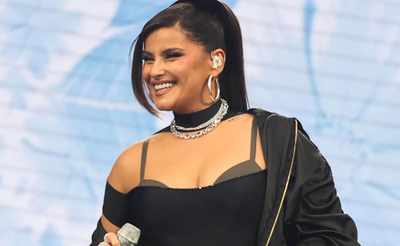 Nelly Furtado zawiesza działalność Uśmiechnięta kobieta ze starannie ułożonymi włosami trzyma mikrofon, ubrana w czarny strój z odkrytymi ramionami i ozdobną biżuterią, na tle jasnej, rozmytej scenerii.