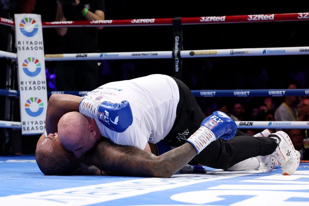 Fabio Wardley w szale radości celebrujący wygraną nad Josephem Parkerem Trener obejmuje leżącego na ringu boksera. Dominują niebiesko-białe rękawice oraz ubrania sportowe, a całość rozgrywa się w otoczeniu ringu z widocznymi reklamami i publicznością.