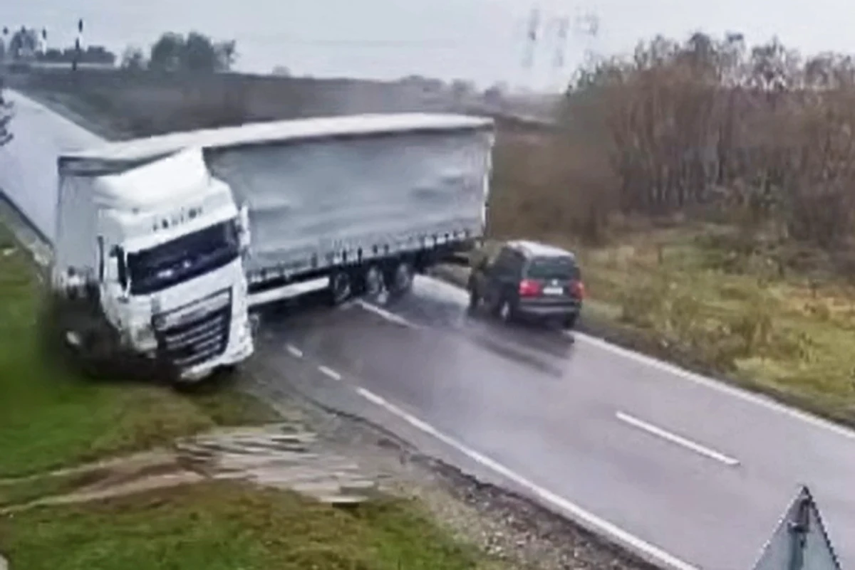 Ciągnik siodłowy z naczepą wpadł w poślizg i stanął w poprzek drogi. Doszło do śmiertelnego wypadku Ciężarówka z naczepą wypadła z jezdni i częściowo zablokowała drogę, zderzając się z samochodem osobowym. Droga jest mokra, otoczona przez trawę i drzewa, panują pochmurne warunki.