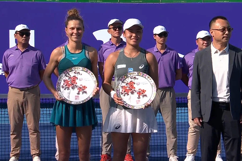 Katarzyna Piter i Janice Tjen z trofeami za triumf w WTA 250 w Kantonie Dwie kobiety tenisistki stoją ramię w ramię na korcie, trzymając okazałe talerze z kwiatowym wzorem jako nagrody. W tle grupa mężczyzn w fioletowych koszulkach oraz jeden mężczyzna w garniturze.