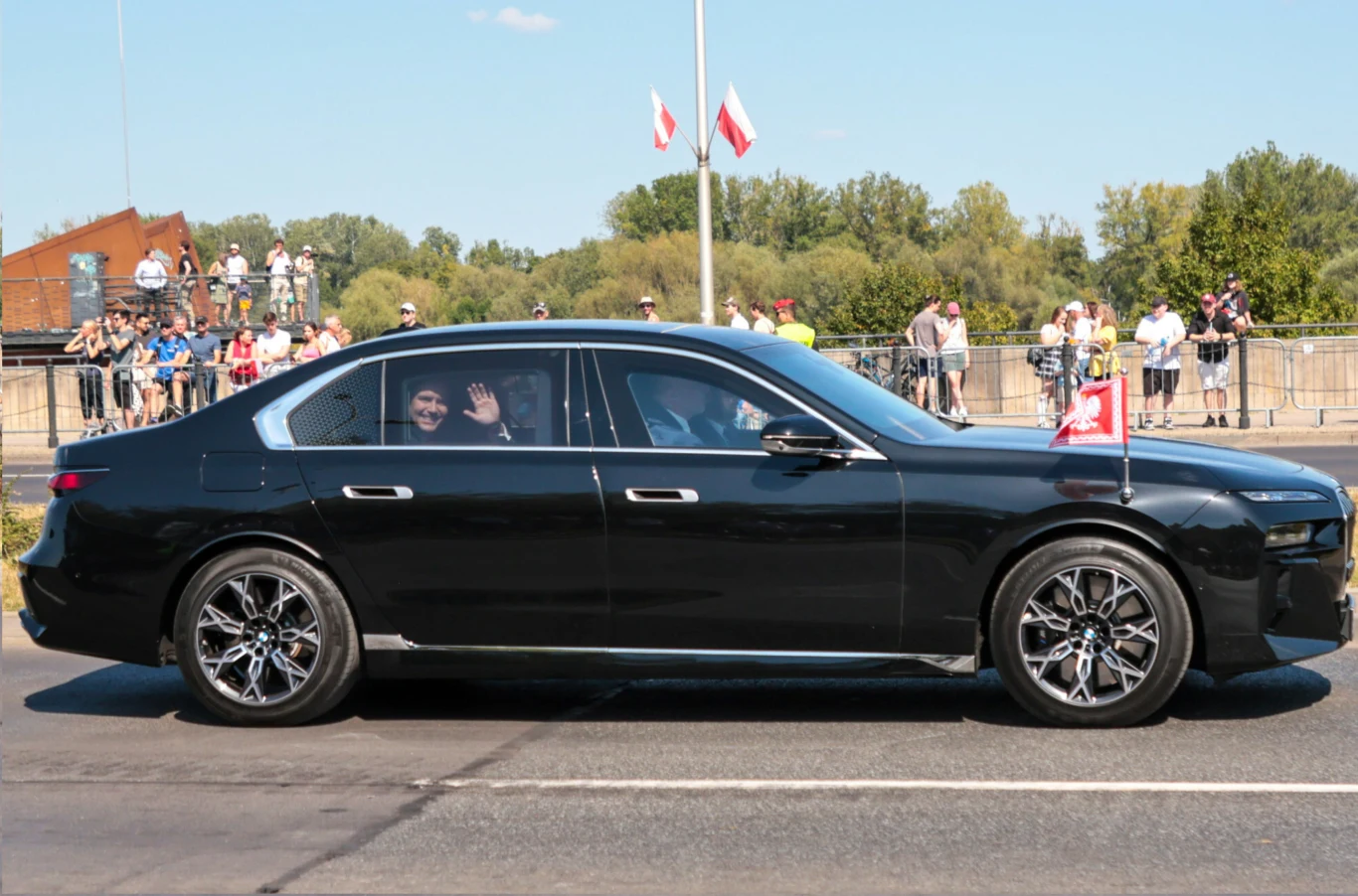 Kancelaria Prezydenta wynajmie osiem limuzyn premium za 3,86 mln zł. Czarny samochód osobowy o charakterze luksusowym przejeżdża ulicą w asyście publiczności stojącej za barierkami, na karoserii pojazdu widoczna jest flaga Polski, w tle drzewa oraz maszt z polską flagą