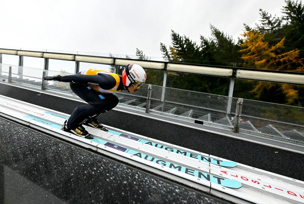 Zawodnik ubrany w kombinezon startowy znajduje się na rozbiegu skoczni narciarskiej, pochylony do przodu w pozycji aerodynamicznej, gotowy do oddania skoku. W tle widoczna jest stalowa konstrukcja oraz drzewa jesienne.