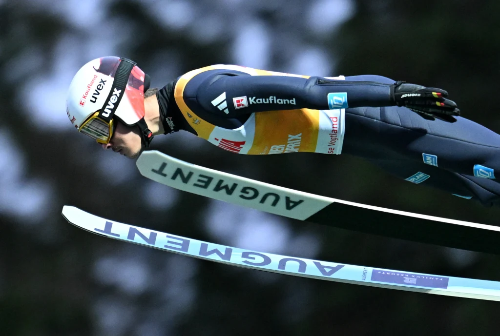 Zawodnik w kombinezonie narciarskim wykonuje skok na nartach podczas zawodów, w tle rozmazane drzewa, wyraźnie widoczne logo sponsorów na stroju oraz nartach.