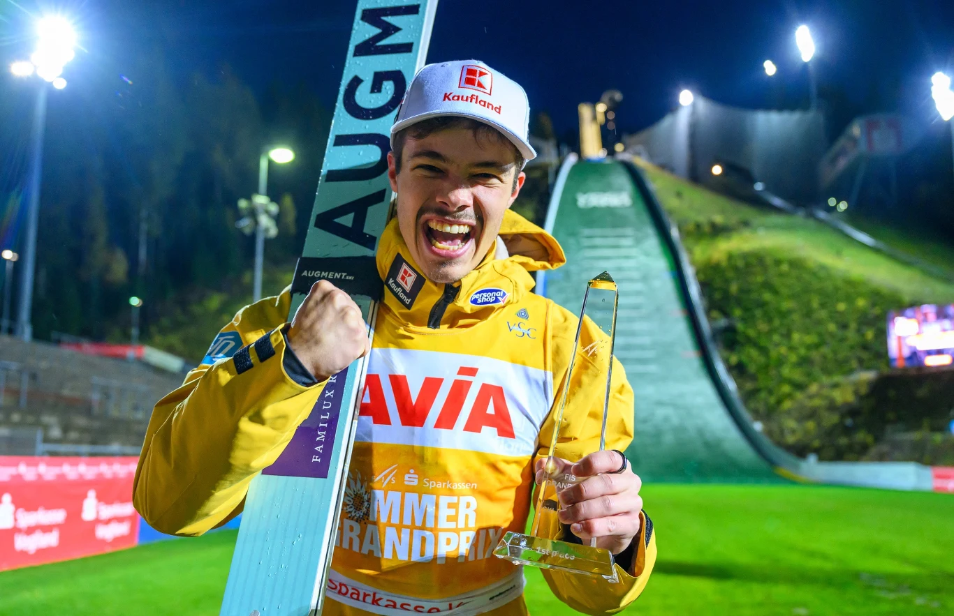 Zwycięski skoczek narciarski w żółtym kombinezonie trzyma w jednej ręce statuetkę, a w drugiej narty, stojąc na tle oświetlonej skoczni narciarskiej podczas nocnych zawodów. Skoczek uśmiecha się szeroko, wyraźnie okazując radość ze zwycięstwa.