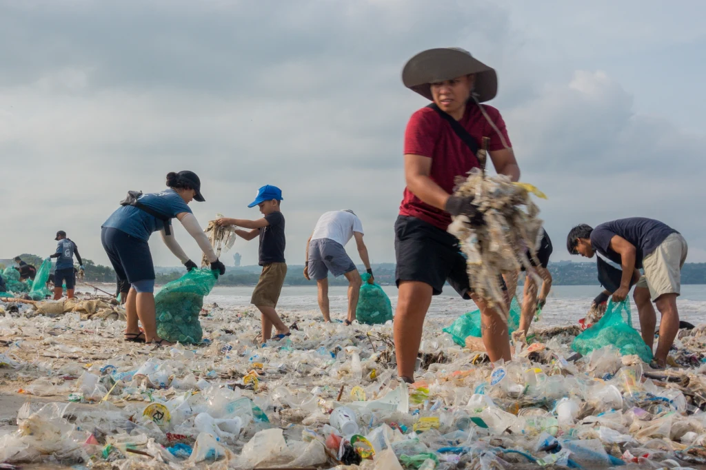 Mikroplastik jest wszechobecny nie tylko w oceanach Grupa ludzi zbiera plastikowe odpady rozrzucone na plaży. W tle morze oraz zachmurzone niebo. Osoby wyposażone są w worki na śmieci, intensywnie pracują, by oczyścić przestrzeń z odpadów.