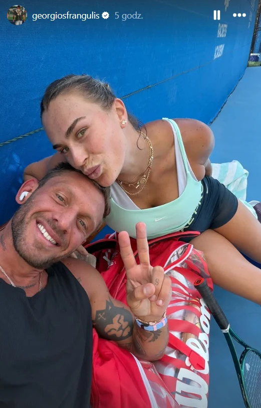 Uśmiechnięty mężczyzna i kobieta pozują razem na tle niebieskiej siatki tenisowej, kobieta siedzi obok dużej czerwonej torby tenisowej i rakiety, mężczyzna pokazuje znak pokoju palcami.