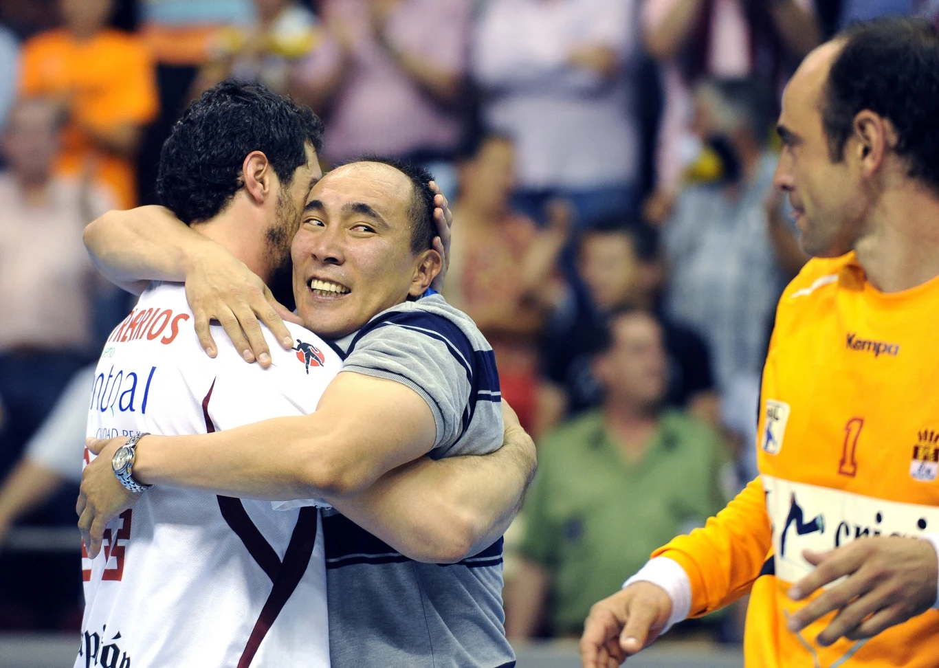 Dwóch mężczyzn obejmuje się, wyrażając radość i emocje, jeden z nich nosi sportową koszulkę, a w tle widać innych uczestników wydarzenia i kibiców.