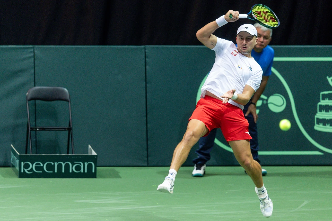 Jan Zieliński rywalizował o deblowy tytuł podczas ATP 500 w Bazylei Zawodnik w białej czapce i czerwonych szortach uderza piłkę tenisową rakietą, będąc w ruchu podczas meczu na korcie, z sędzią w tle oraz pustym krzesłem przy bandzie reklamowej.