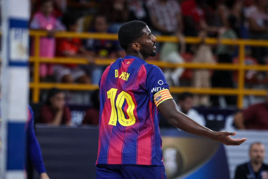 Piłkarz w koszulce z numerem 10 i nazwiskiem na plecach, grający dla drużyny w barwach bordowo-granatowych podczas meczu na hali, w tle widoczni kibice oraz żółte barierki.