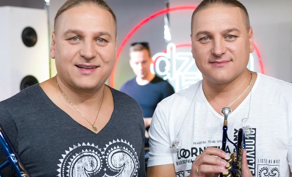 Dwóch mężczyzn o jasnych włosach i podobnym wyglądzie, stojących obok siebie z instrumentami muzycznymi w dłoniach. W tle rozmyta postać oraz okrągły neon.