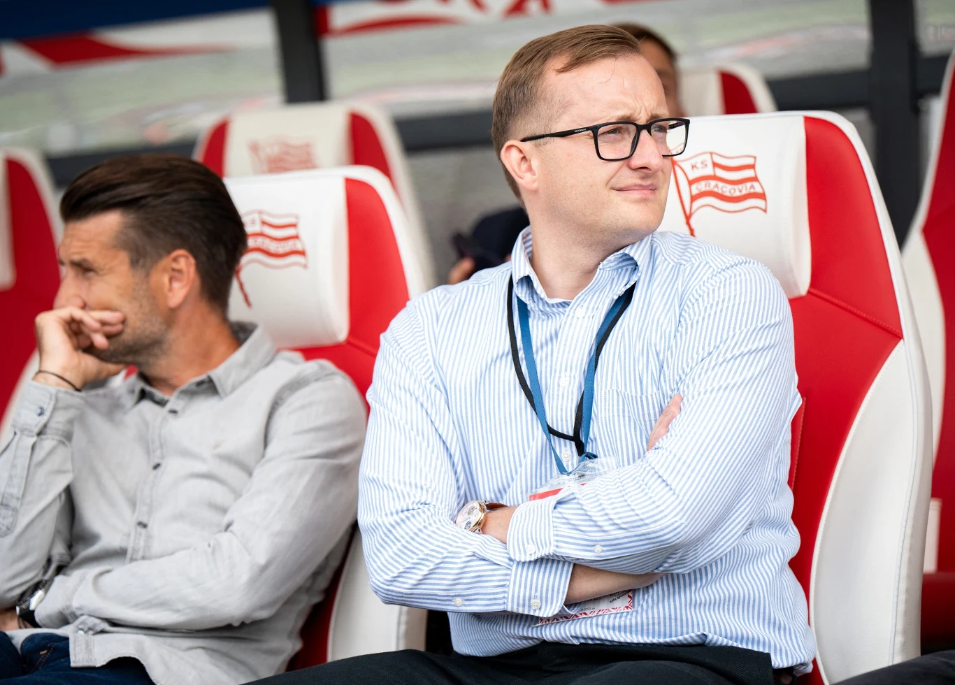 Dwóch mężczyzn siedzących na stadionowych fotelach z logotypem klubu, jeden w okularach, w koszuli i z identyfikatorem na szyi, drugi ubrany bardziej swobodnie, obaj skupieni i obserwujący coś poza kadrem.