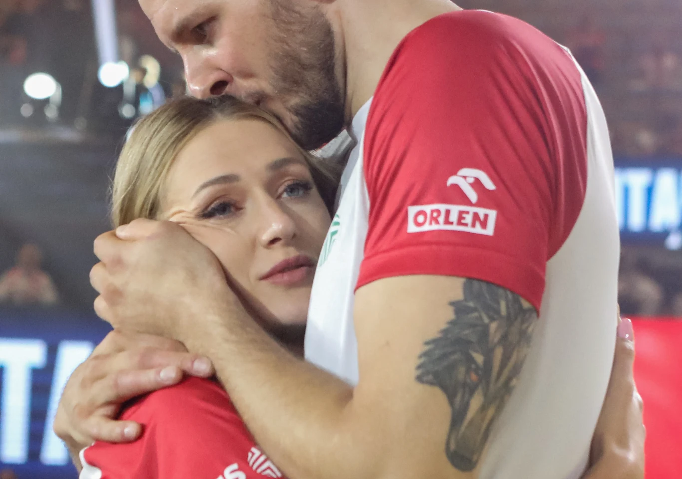 Mężczyzna w sportowej koszulce przytula i całuje na głowie kobietę, która wygląda na wzruszoną lub smutną, tworząc emocjonalny i intymny moment.