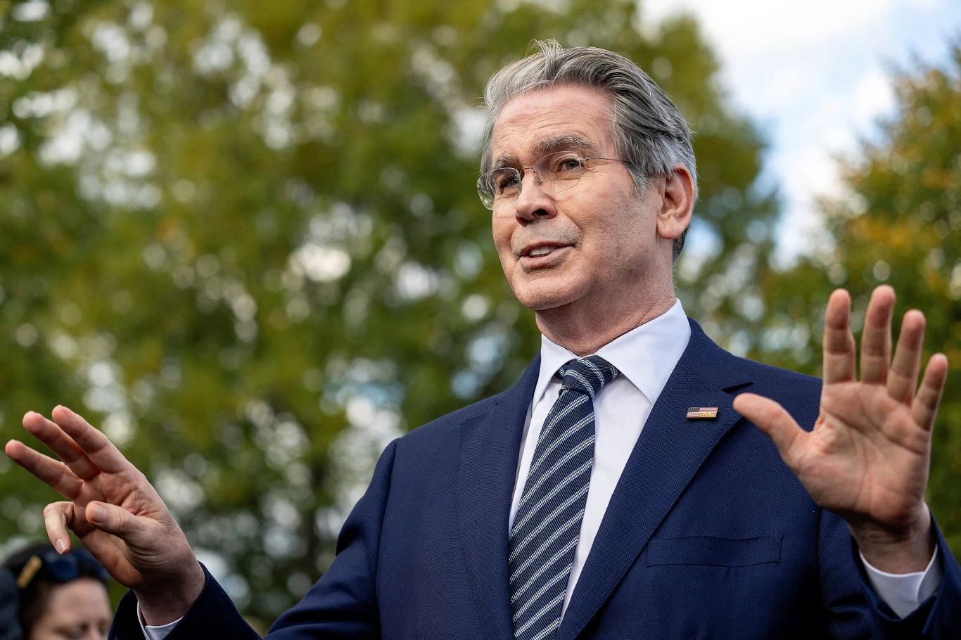 Minister finansów USA Scott Bessent Mężczyzna w średnim wieku w garniturze, z siwymi włosami, trzyma uniesione dłonie w geście podkreślającym wypowiedź, w tle zielone drzewa i delikatnie rozmazane sylwetki innych osób.