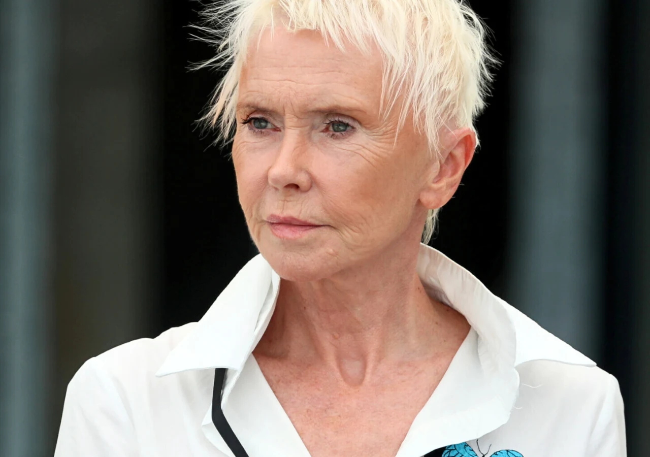 Ewa Błaszczyk Starsza kobieta o krótkich, jasnych włosach patrzy przed siebie, ubrana w białą koszulę z wywiniętym kołnierzem, na tle rozmazanego ciemnego tła.