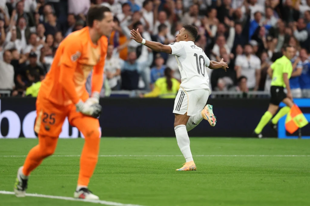 Szczęsny pokonany przez Mbappe Jeden piłkarz w białym stroju z numerem 10 unosi ręce w geście triumfu, podczas gdy bramkarz w pomarańczowym stroju wydaje się zrezygnowany. W tle sędzia oraz wiwatujący tłum na trybunach.